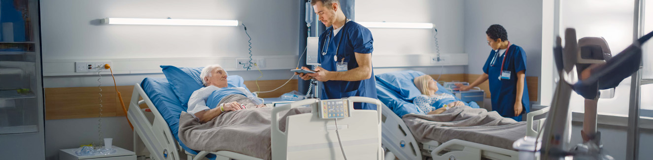 Hospital Ward Friendly Male Nurse Talks Reassuringly to Elderly Patient Resting in Bed